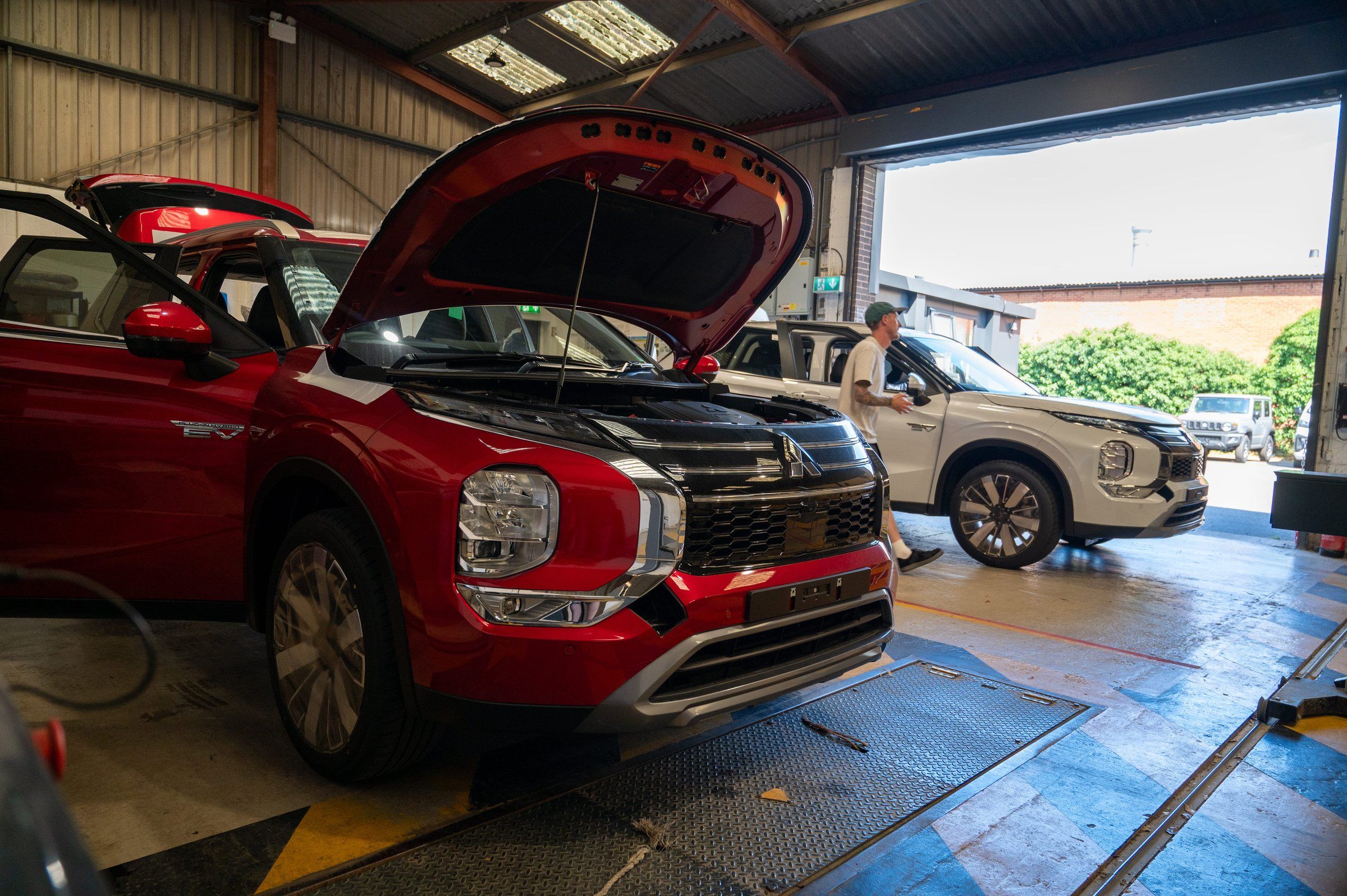 A red Mitsubishi Outlander plug-in hybrid with its bonnet up on the inspection lane in the My Car Import workshop